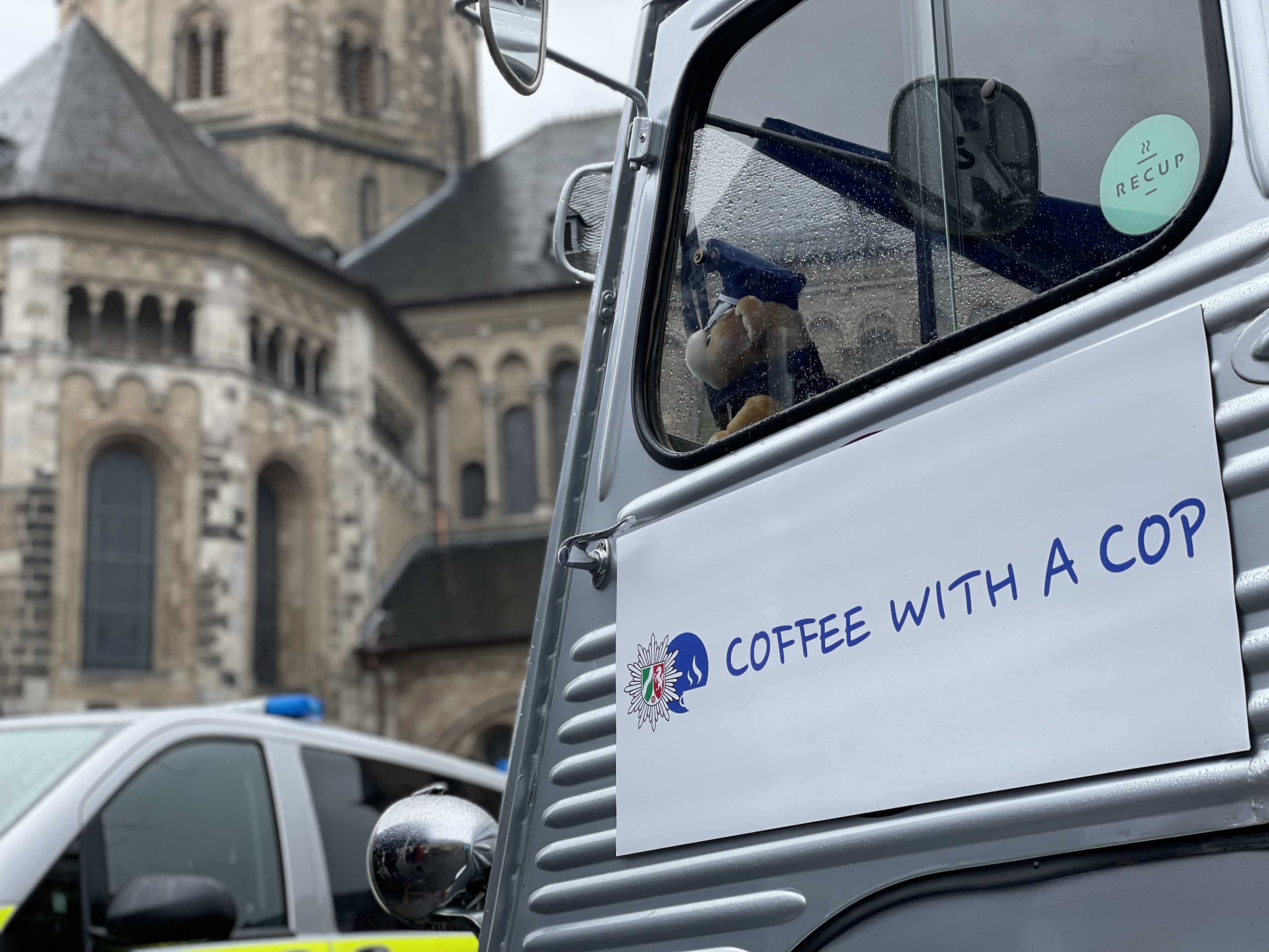 Logo der Aktion "Coffee with a Cop" auf einer Seitentür. Das Logo zeigt den Polizeistern und eine Tasse Kaffee in einem blauen Kreis sowie den Schriftzug Coffee with a Cop. Im Hintergrund ein Streifenwagen der Polizei und die Bonner Münsterkirche
